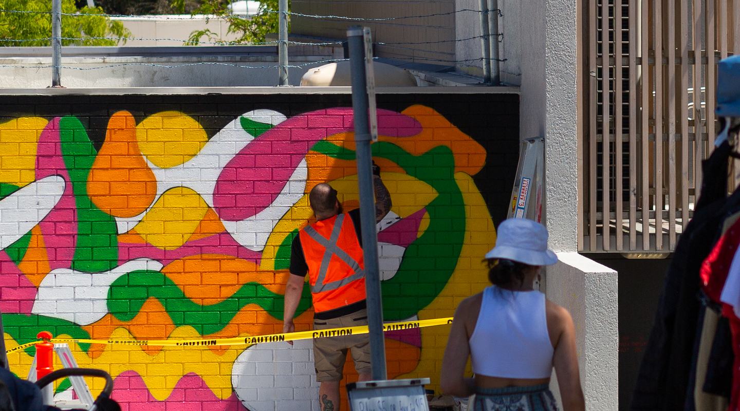 A crowd gathers in the shade of a grey barked gumtree to watch Paul Eggins work on his mural of yellow, pink and green abstract shapes. In the foreground is a market stall tent displaying second hand clothing for sale.
