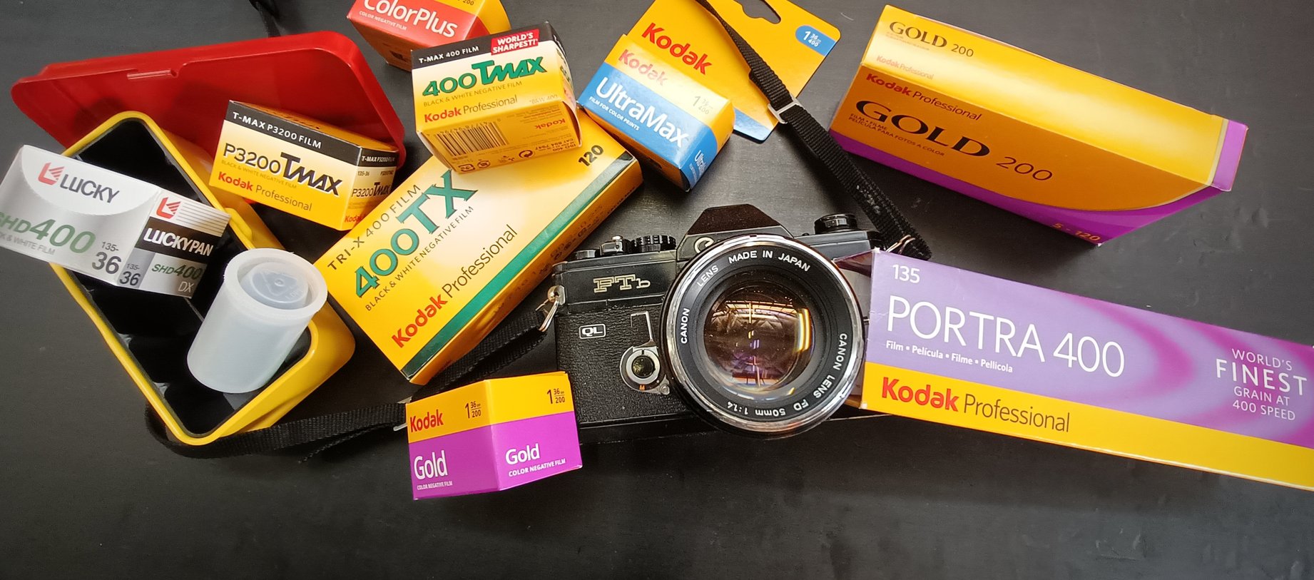 Photograph of vibrant yellow boxes of film scattered around a Canon film camera in front of a black background