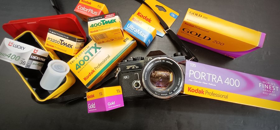 Photograph of vibrant yellow boxes of film scattered around a Canon film camera in front of a black background
